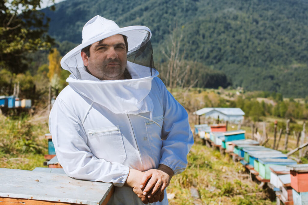 Imker David in een wit imkerpak, zijn bijenkorven op de achtergrond