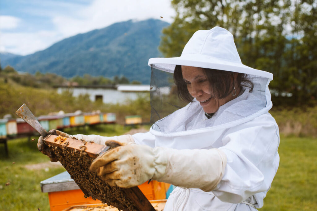 Imker Carmen in een wit imkerpak glimlacht en kijkt naar een honingraat waarop veel bijen zitten
