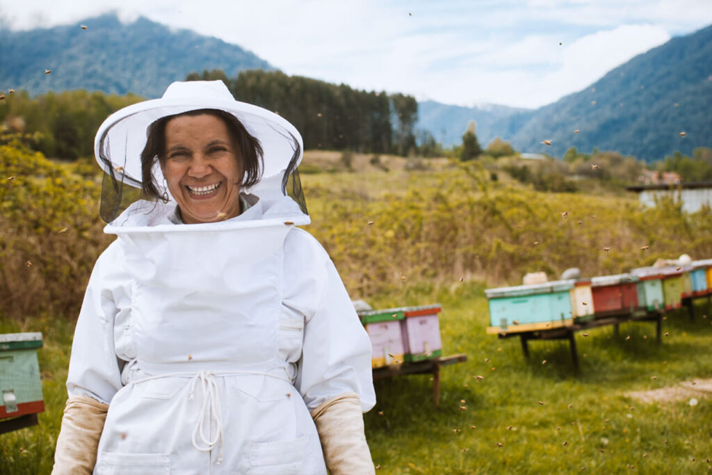 Glimlachende imker Carmen in een wit imkerpak voor haar kleurrijke bijenkorven