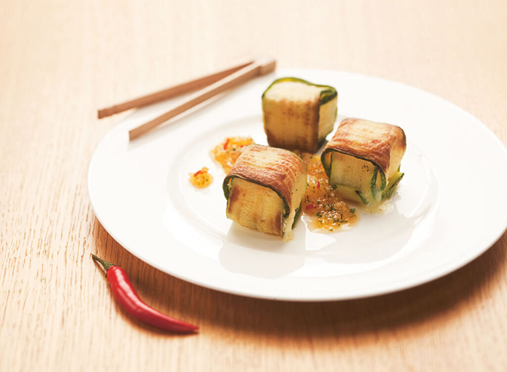 Drie in courgette verpakte kaaspakketjes op een wit bord. Een rode chilipeper voor het bord