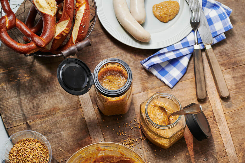 De tafel is gedekt met een mandje pretzels, een bord met twee witte worstjes en een klodder mosterd. Twee glazen zoete mosterd en een schaaltje mosterdzaadjes