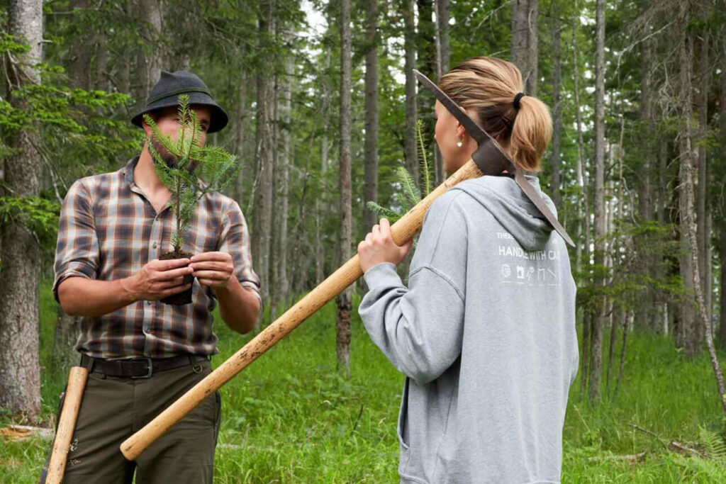 Twee mensen praten, de vrouw heeft een schoffel over haar schouder, de man een zaailing in zijn hand. Op de achtergrond is een bos