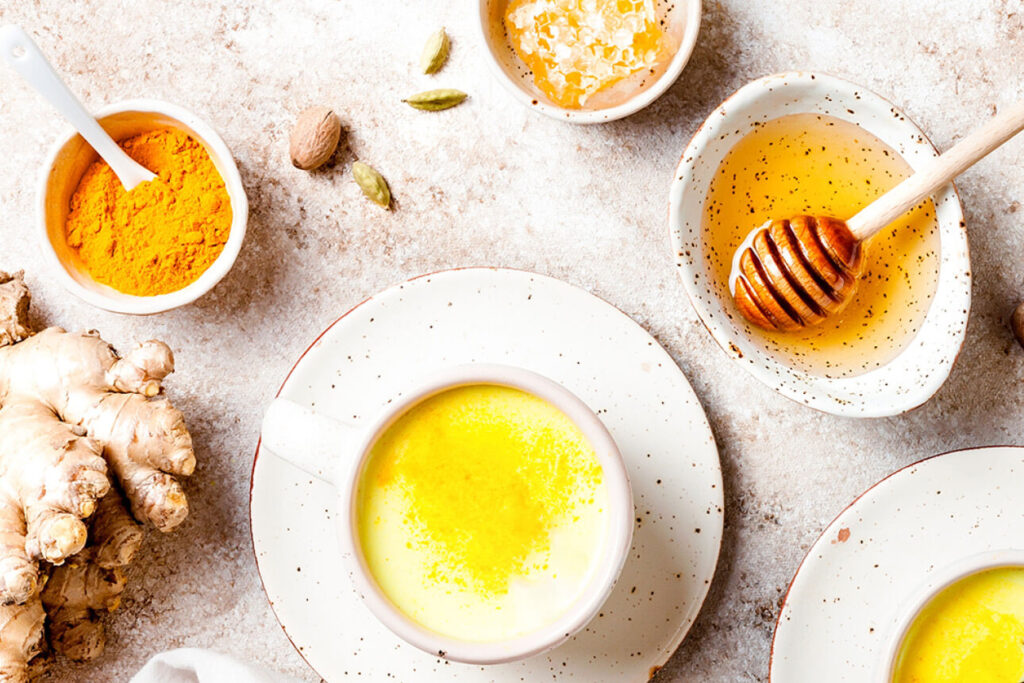 Bovenaanzicht van een beker gevuld met honingmelk. Ernaast staat een schaaltje honing met een honinglepel erin. Er ligt ook een gemberwortel op tafel