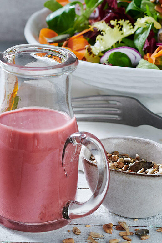 Karaf met honing-mosterd dressing naast een kom geroosterde zaden, op de achtergrond een bord met een kleurrijke gemengde salade